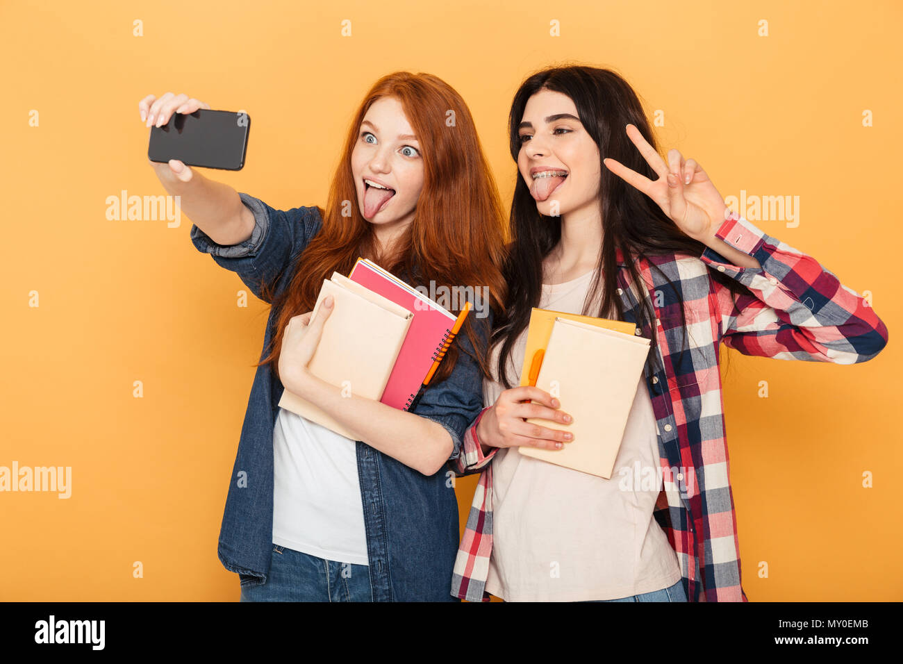 Portrait de deux drôles de jeunes adolescentes de l'école tenue des cahiers et en tenant un isolé sur fond jaune selfies Banque D'Images