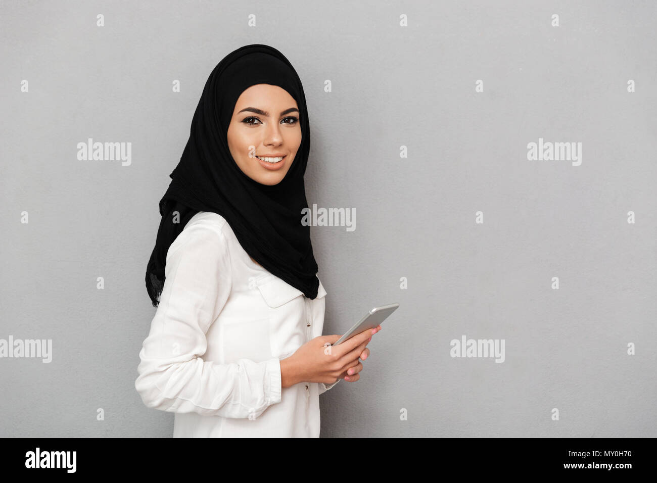 Superbe Portrait de femme arabe en voile avec maquillage oriental holding silver smartphone pendant à la caméra à l'arrière-plan gris plus isolés Banque D'Images