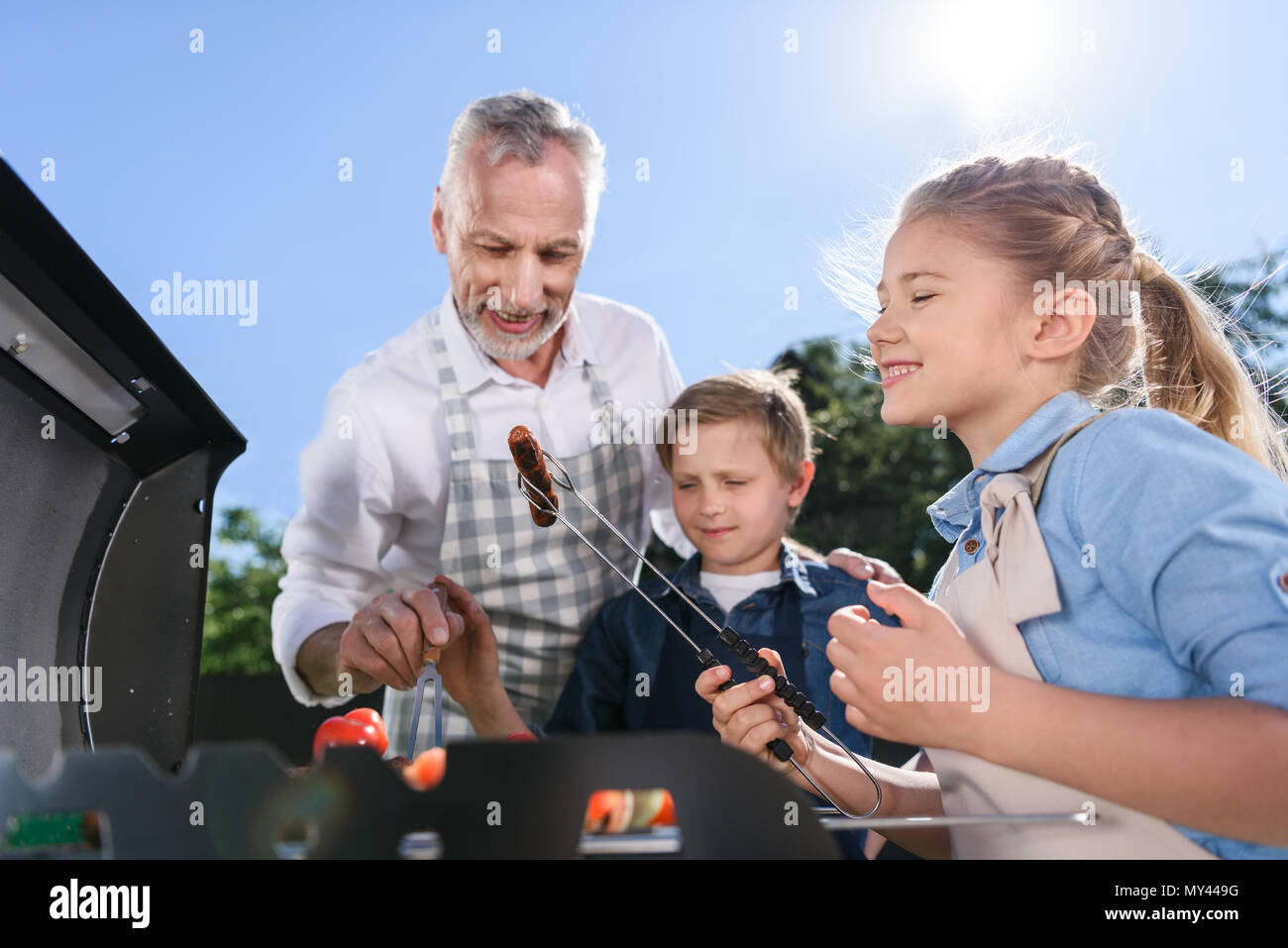 Petits-enfants avec grand-père préparer des saucisses sur le gril du barbecue en plein air Banque D'Images