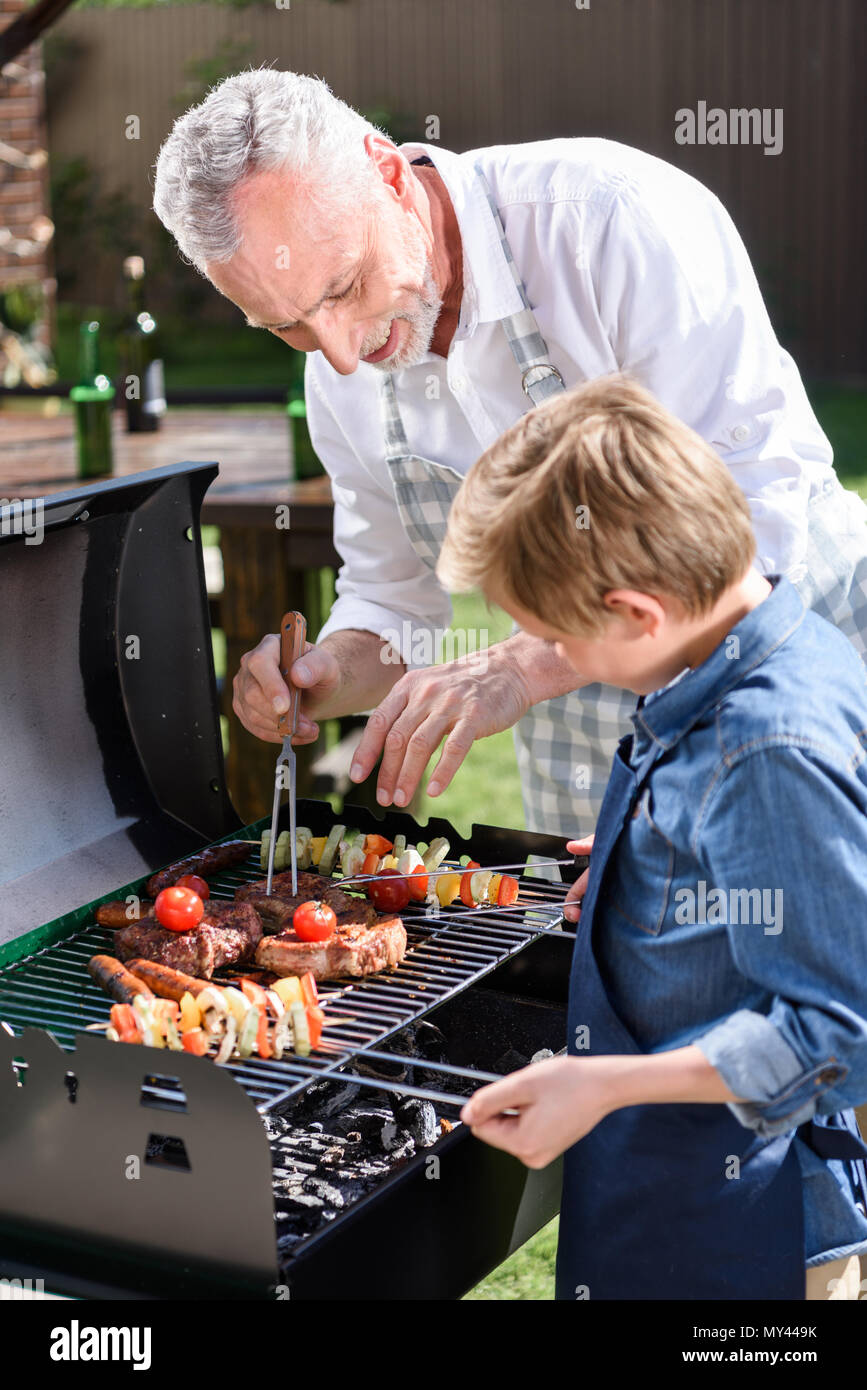 Grand-père aux cheveux gris avec son petit-fils préparer la viande et les légumes sur le gril en plein air Banque D'Images