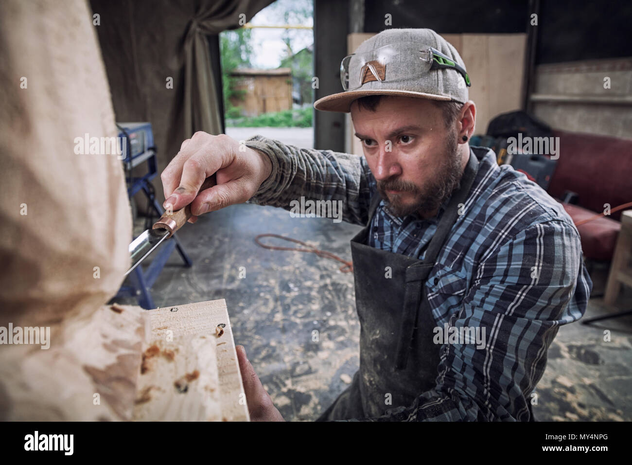 Jeune homme dans un menuisier scies vêtements de travail à la tête d'un homme avec un arbre , à l'aide d'un burin dans l'atelier, autour d'un lot d'outils pour le travail Banque D'Images