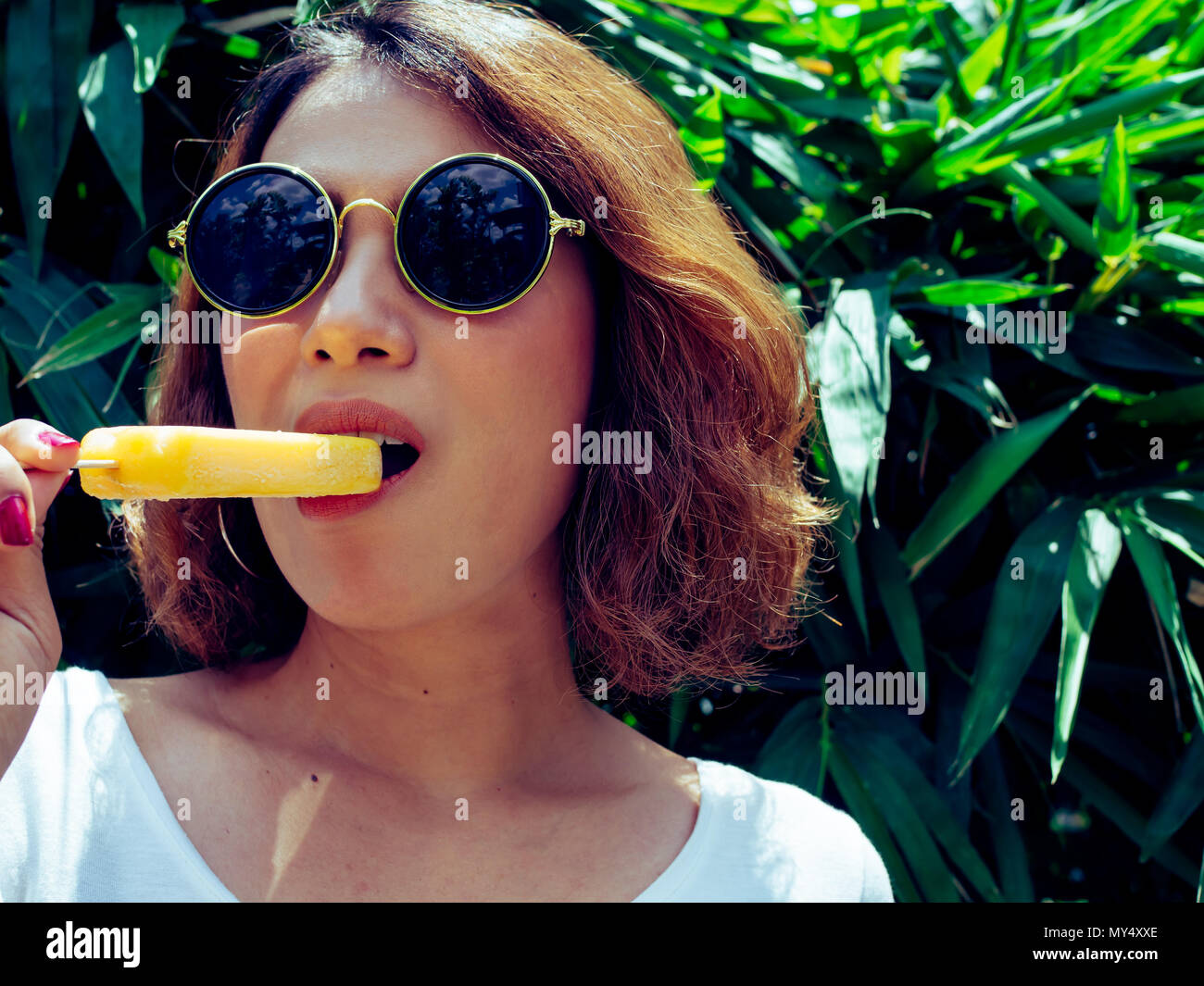 Belle femme cheveux courts portant chemise blanche et lunettes ronde jaune  manger ma glace Ice Melting Pop sur l'heure d'été, avec du soleil sur la  zone de l'arbre Photo Stock - Alamy