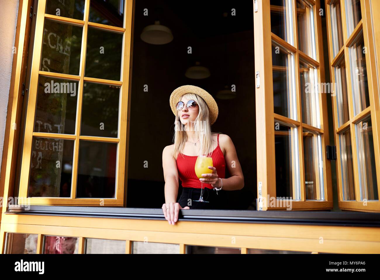 Une jeune blonde est sympathique, dans un café près d'une fenêtre ouverte et est titulaire d'un verre de cocktail. Banque D'Images