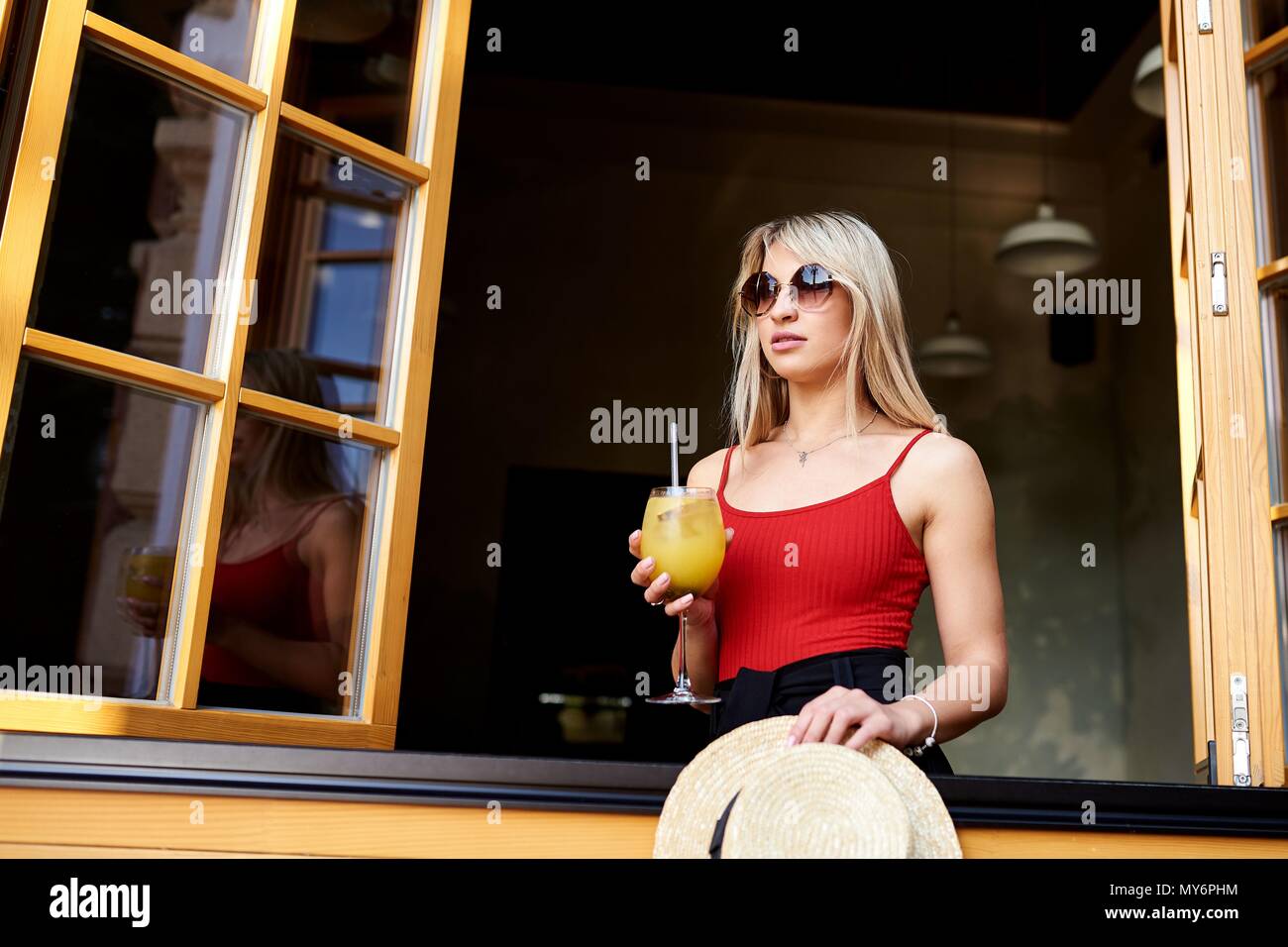 Une jeune blonde est sympathique, dans un café près d'une fenêtre ouverte et est titulaire d'un verre de cocktail. Banque D'Images
