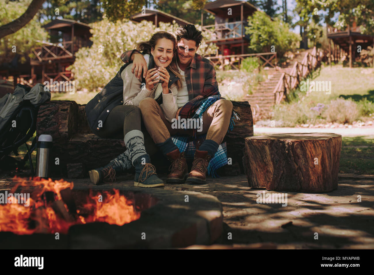 Heureux jeune homme et femme assise par camp avec du café. Couple aimant se reposer par un feu de camping. Banque D'Images