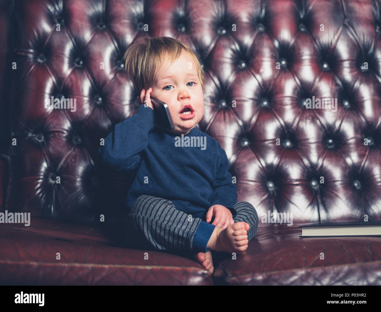 Un mignon petit bébé garçon est assis sur un canapé en cuir et est en train de parler sur un téléphone intelligent Banque D'Images