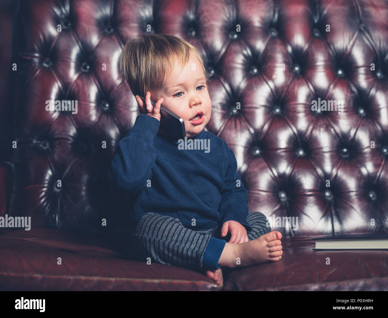 Un mignon petit bébé garçon est assis sur un canapé en cuir et est en train de parler sur un téléphone intelligent Banque D'Images