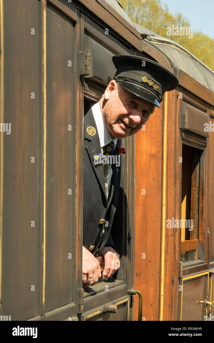 Close up Portrait de gentilhomme contrôleur sur Severn Valley Railway, penché hors de la fenêtre transport au soleil, prêt à accueillir les passagers. Banque D'Images