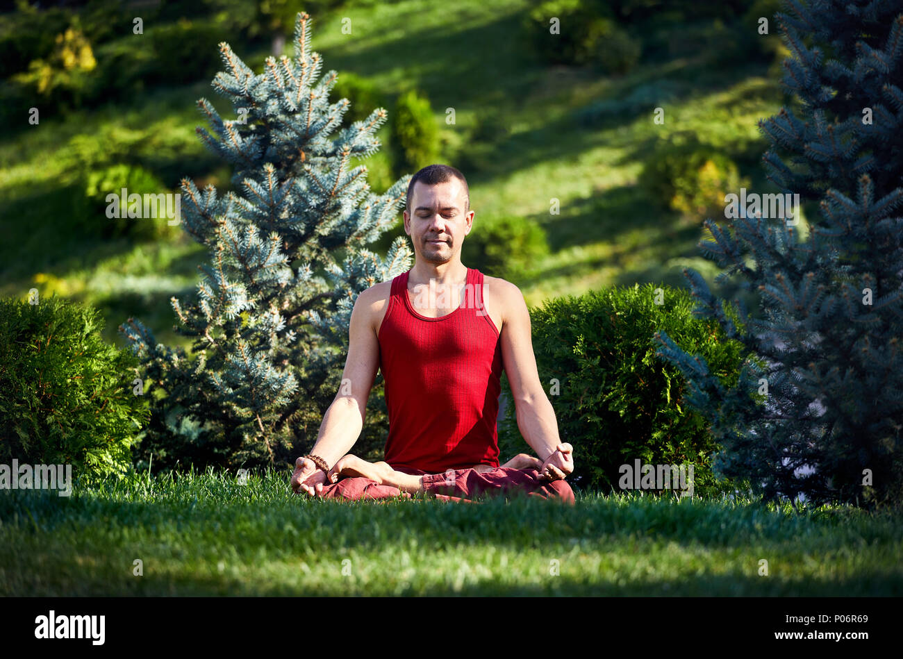 Beau mettre en place l'homme en chemise rouge est de méditer sur l'herbe verte dans le parc avec les yeux fermés le jour d'été ensoleillé. Concept de la méditation et sain lifesty Banque D'Images
