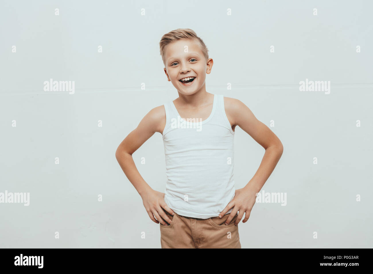 Young smiling boy en débardeur blanc debout avec les mains sur les hanches et looking at camera, isolated on white Banque D'Images