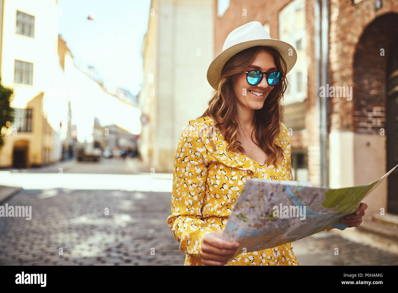 Souriante jeune femme brune portant un chapeau et des lunettes à explorer la ville avec une carte Banque D'Images