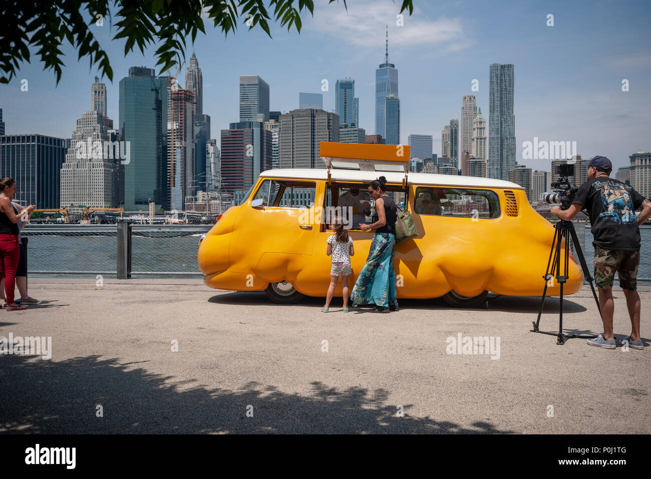 Brooklyn, Etats-Unis. 9 juin 2018. Avec le ciel de New York derrière eux les visiteurs au parc Du Pont de Brooklyn à New York delight dans artiste Erwin Wurm's 'Hot Dog' de bus desservant gratuitement des hot-dogs à toute et tous, vu le jour de l'ouverture, samedi, Juin 9, 2018. L'artiste autrichien modifié un vintage Volkswagen Microbus dans un camion alimentaire jaune vif à bulbe, redéfinir la dichotomie entre commerce et la sculpture, bien qu'emblématique, où vous profiterez gratuitement de l'alimentation de rue de New York. Le chariot était auparavant le 'BUS' Curry wursts servant en Europe. Crédit : Richard Levine/Alamy Live News Banque D'Images