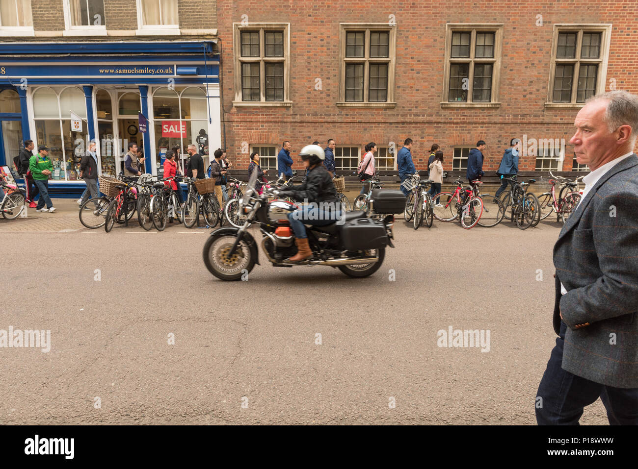 Le motocycliste passe devant les piétons et les vélos garés dans une rue animée de Cambridge, capturant la vie urbaine, les mouvements et les déplacements en ville au Royaume-Uni. Banque D'Images