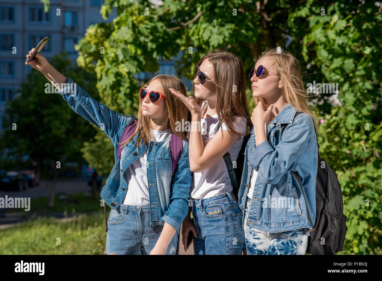 Trois adolescentes en été à l'extérieur. Fait une photo de la personne sur  le téléphone. Ils portent des jeans élégant vêtements. Des lunettes de  soleil. Les adolescentes sont blondes avec des sacs