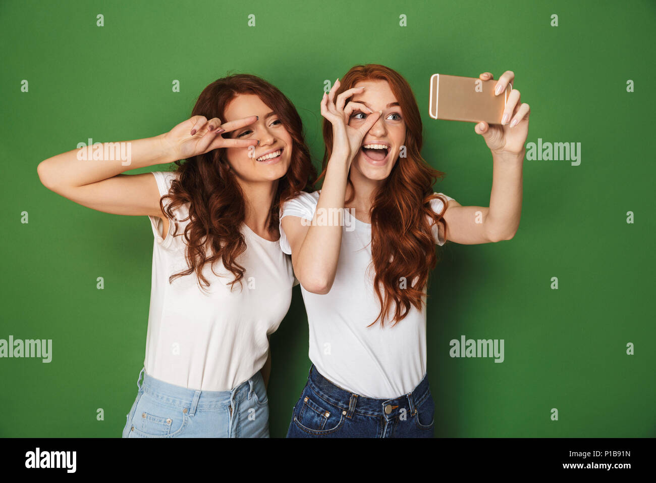 Image de deux adolescentes drôle avec ginger hair montrant ok et signe de la paix près de yeux tout en tenant le téléphone cellulaire selfies sur fond vert isolé Banque D'Images