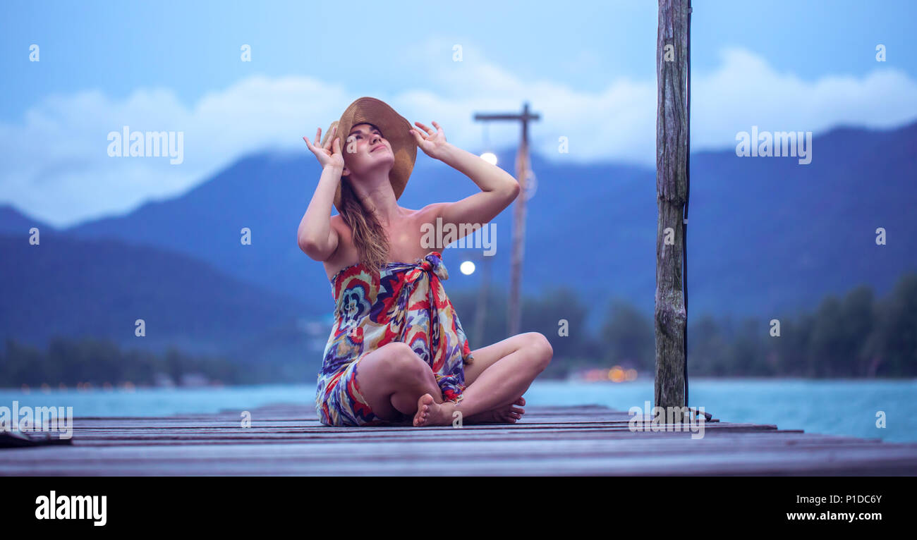 Belle fille dans un chapeau assis sur une longue jetée en bois dans la soirée sur Koh Chang, Thaïlande Banque D'Images