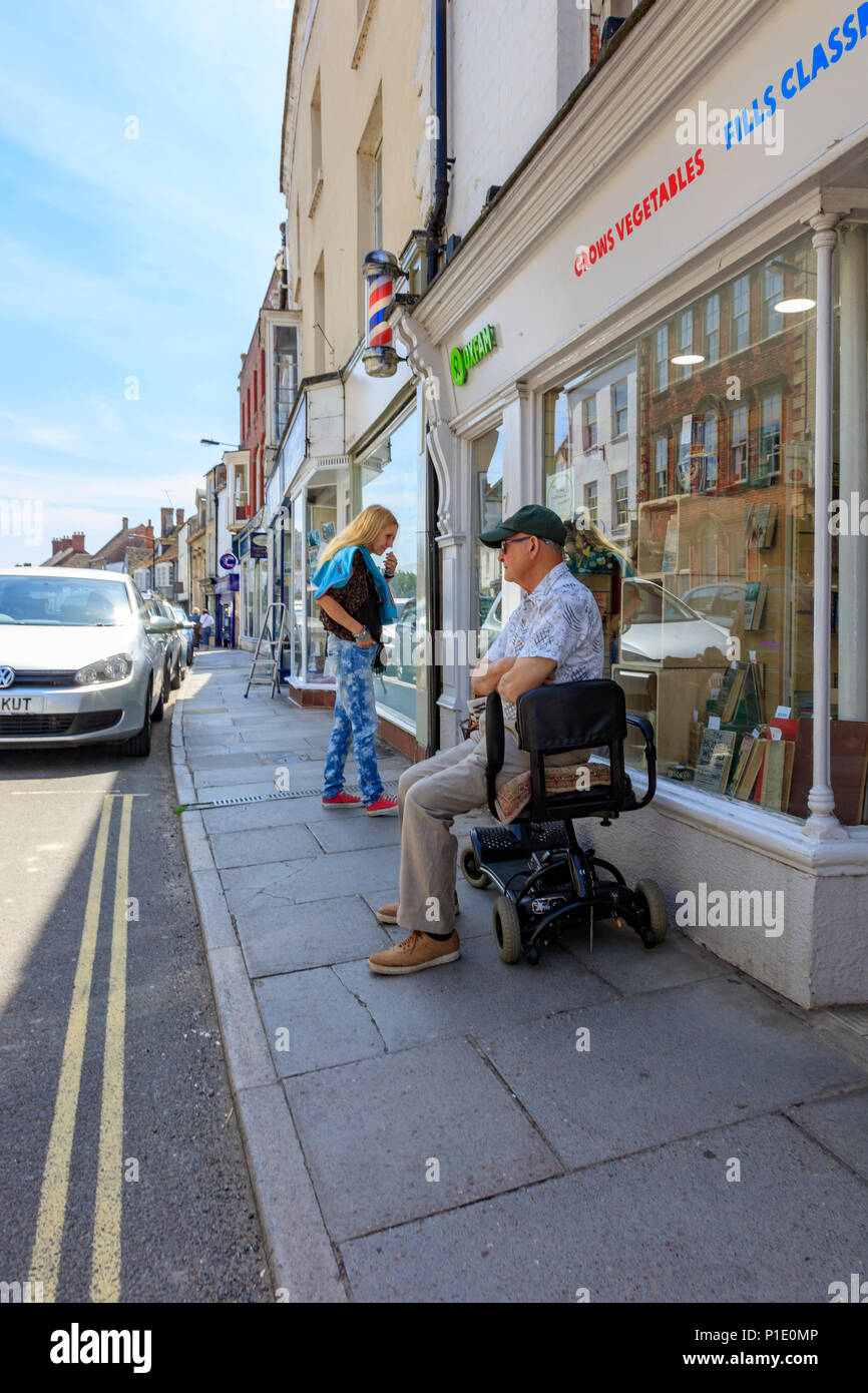 Un homme est assis sur un fauteuil roulant pendant qu'il attend de sa partenaire qui est dans le magasin de charité Oxfam, Malmesbury High Street, Wiltshire, Royaume-Uni Banque D'Images