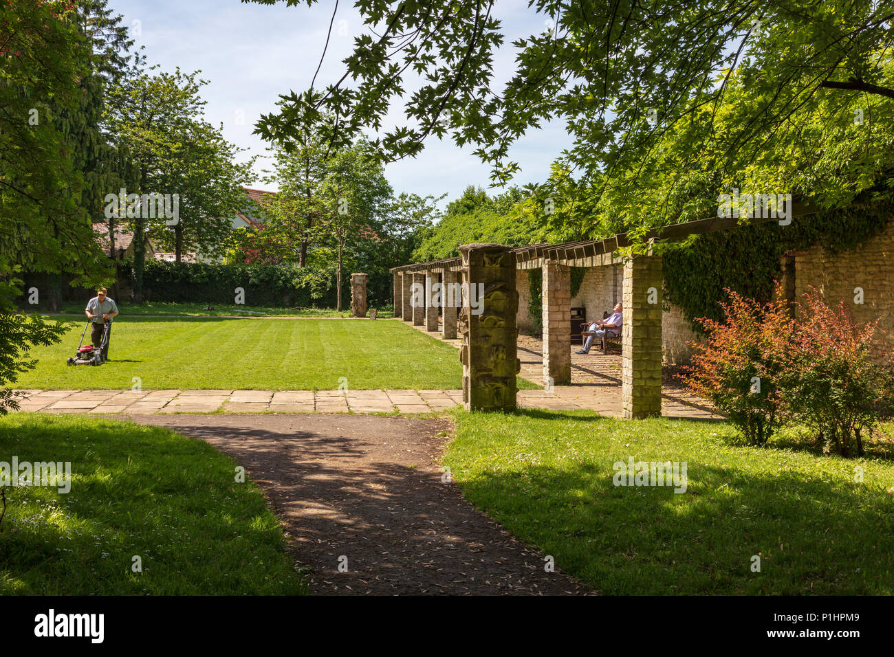 Un jardinier qui tond la pelouse à Malmesbury Abbey Gardens, alors qu'un visiteur se trouve dans le soleil et des silences, Wiltshire, UK Banque D'Images