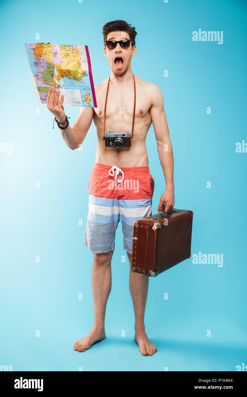 Portrait d'un jeune homme torse nu en a choqué les caleçons de bains tenant  une carte tout en exerçant son appareil photo et photo valise sur fond bleu  Photo Stock - Alamy