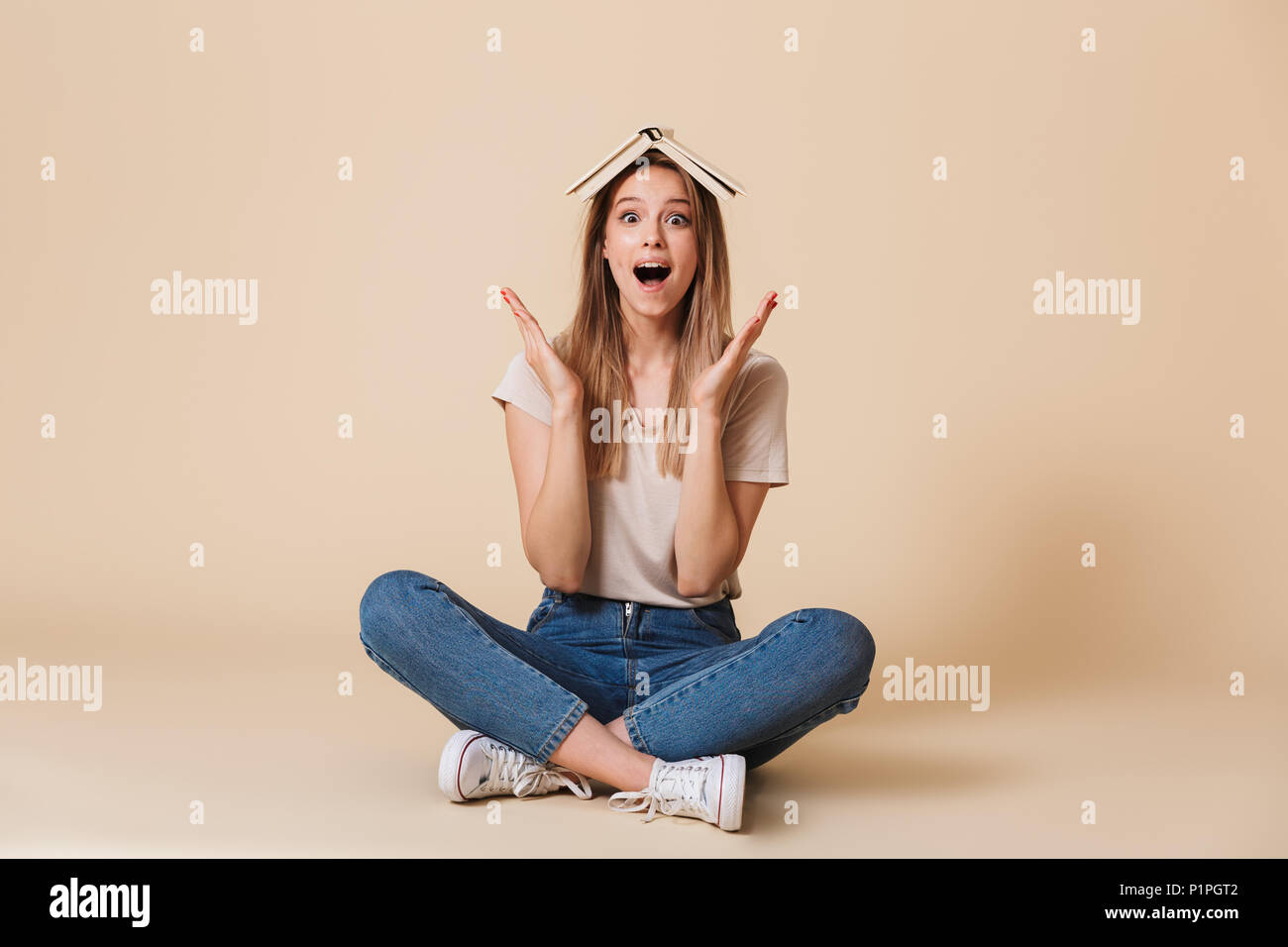 Photo de amusant drôle femme portant des vêtements décontractés assis au sol avec les jambes croisées et livre ouvert sur fond beige plus isolé de la tête Banque D'Images