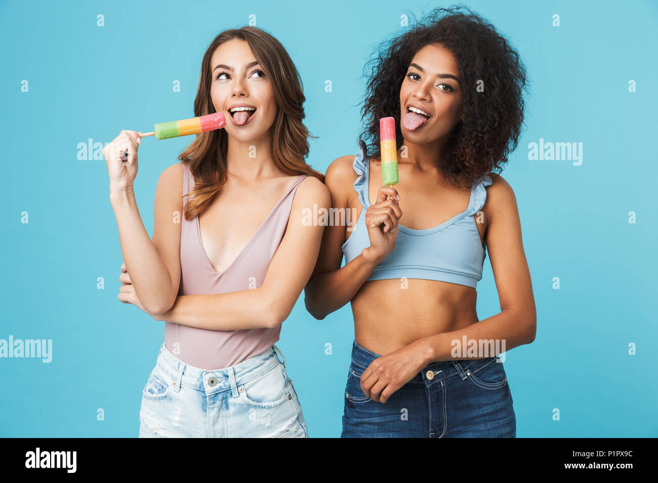 Deux jolies jeunes filles habillés en vêtements d'été de lécher la crème glacée isolé sur fond bleu Banque D'Images