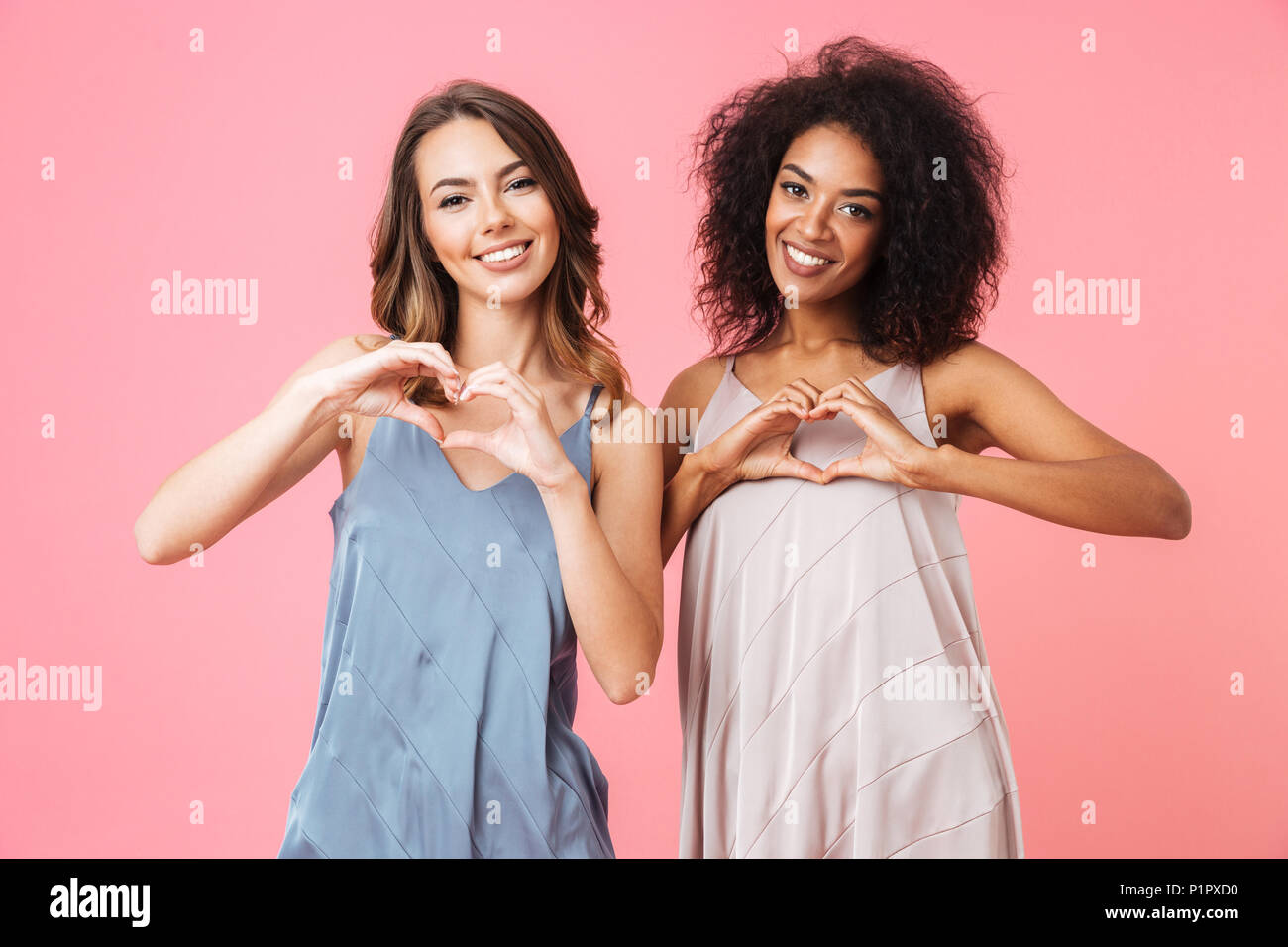 Deux jeunes filles gaie habillés en vêtements d'été montrant coeur geste avec les doigts plus isolé sur fond rose Banque D'Images