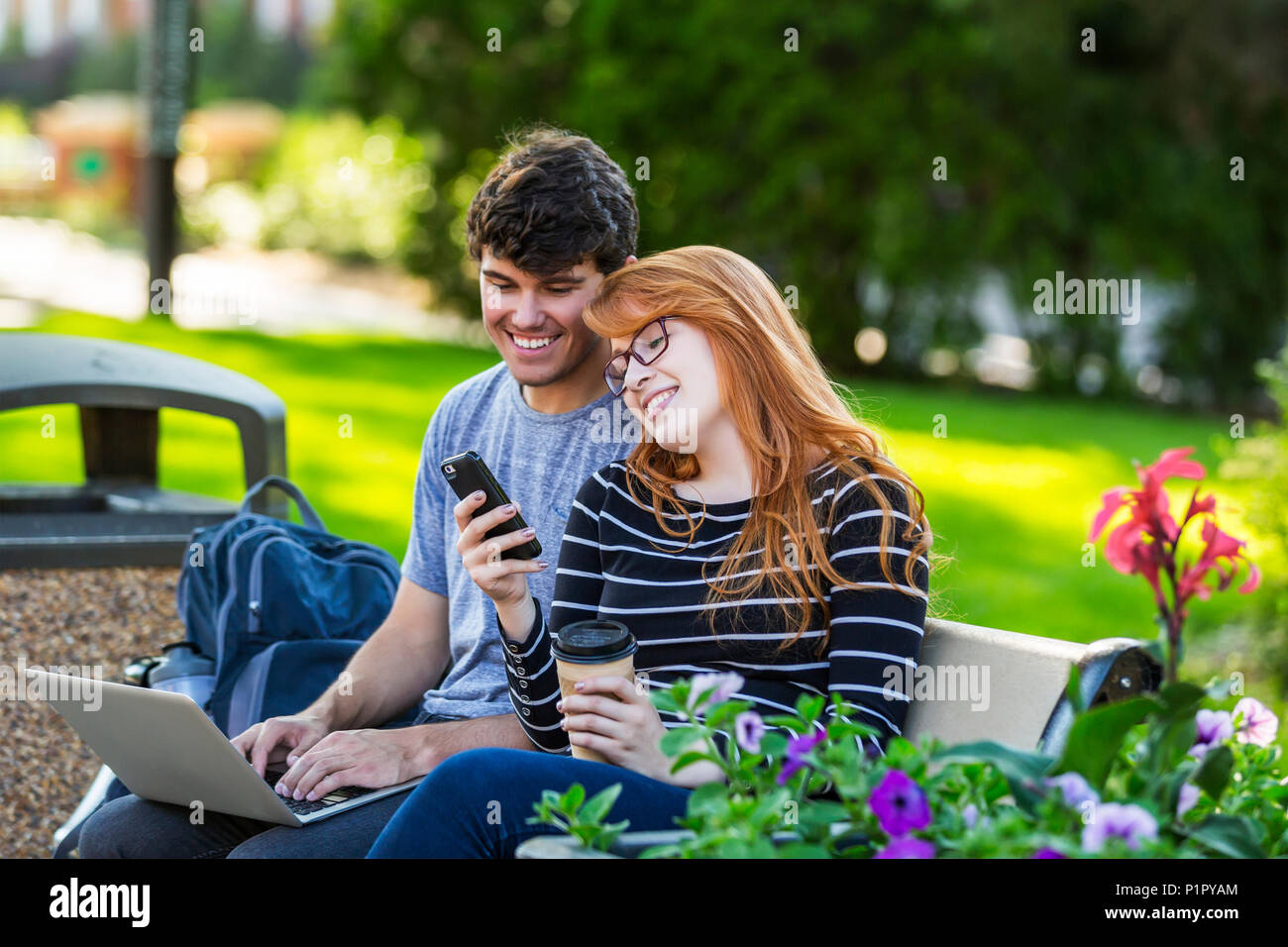 Un jeune homme et une jeune femme s'assoient sur un banc sur le campus universitaire à l'aide d'un ordinateur portable et vérifient les médias sociaux sur un smartphone tout en... Banque D'Images