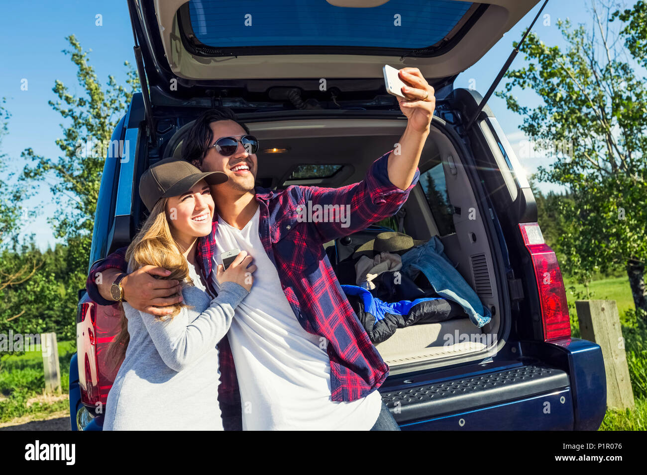 Jeune couple à leur véhicule avec le dos ouvert en tenant un auto-portrait avec leur téléphone cellulaire ; Edmonton, Alberta, Canada Banque D'Images