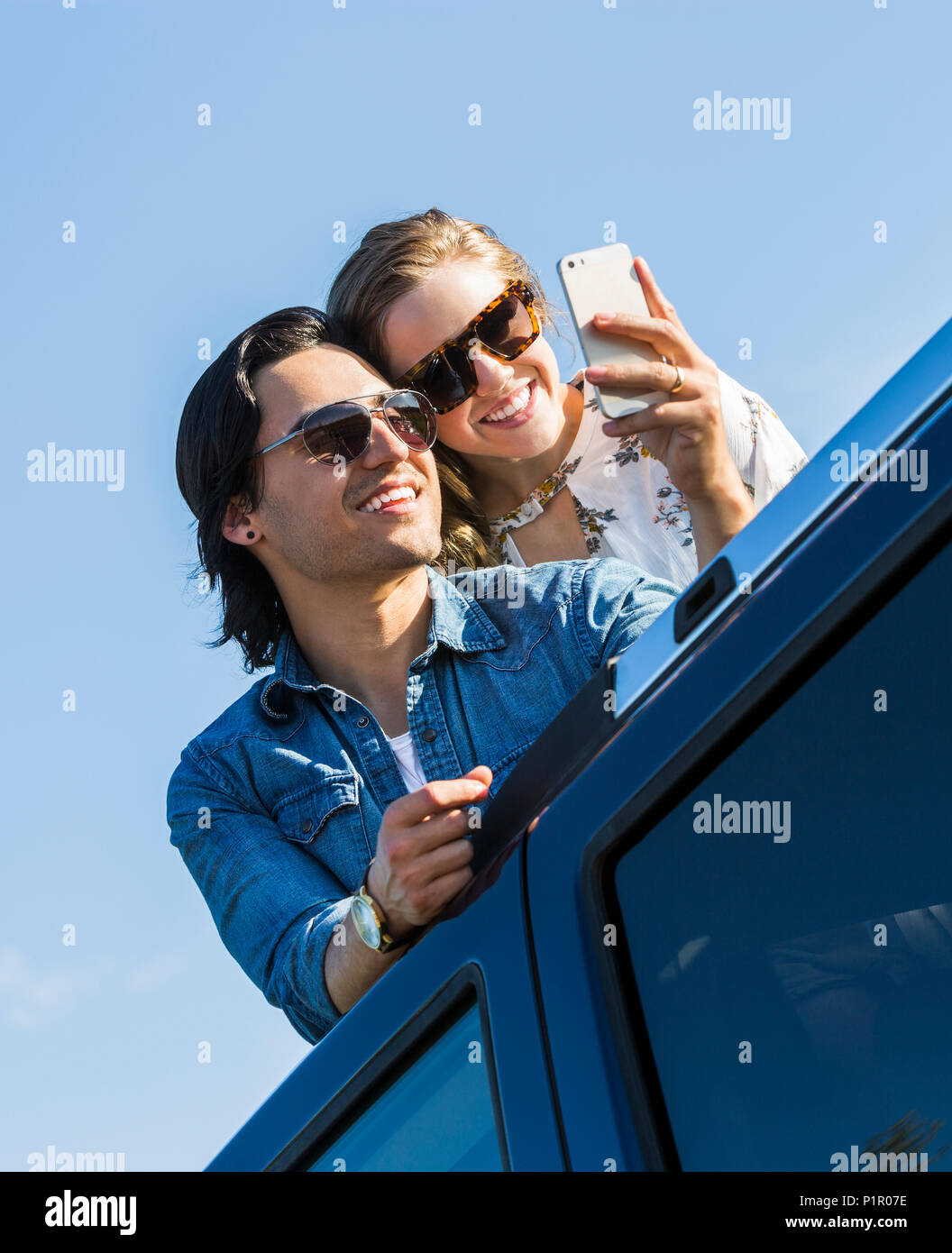 Un jeune couple debout à travers le toit ouvrant de leur véhicule et de faire un autoportrait avec leur téléphone cellulaire ; Edmonton, Alberta, Canada Banque D'Images