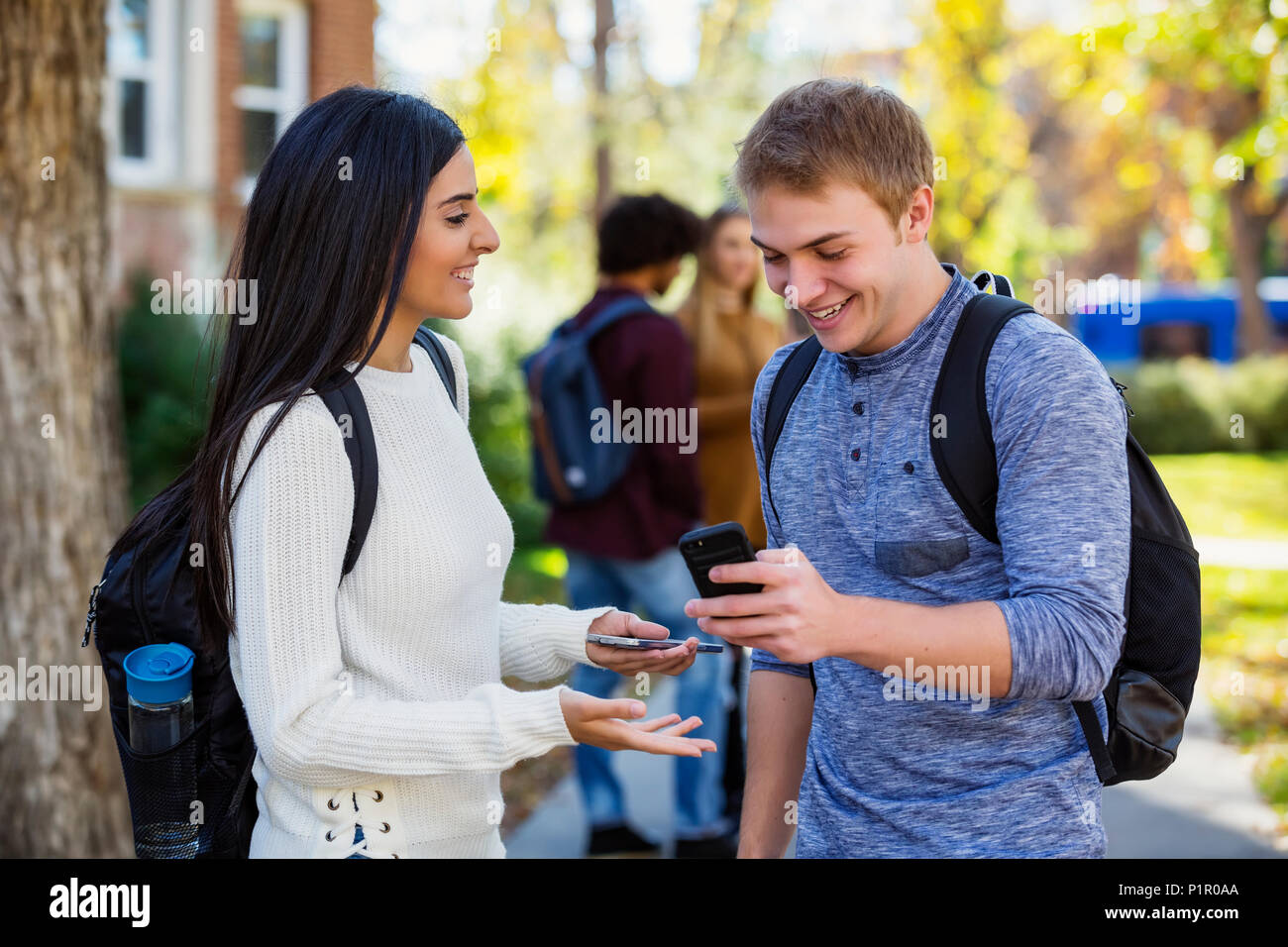 Un étudiant d'université mâle et femelle se trouve sur un chemin regardant un smartphone ensemble, riant et parlant avec un petit groupe de confrères ... Banque D'Images