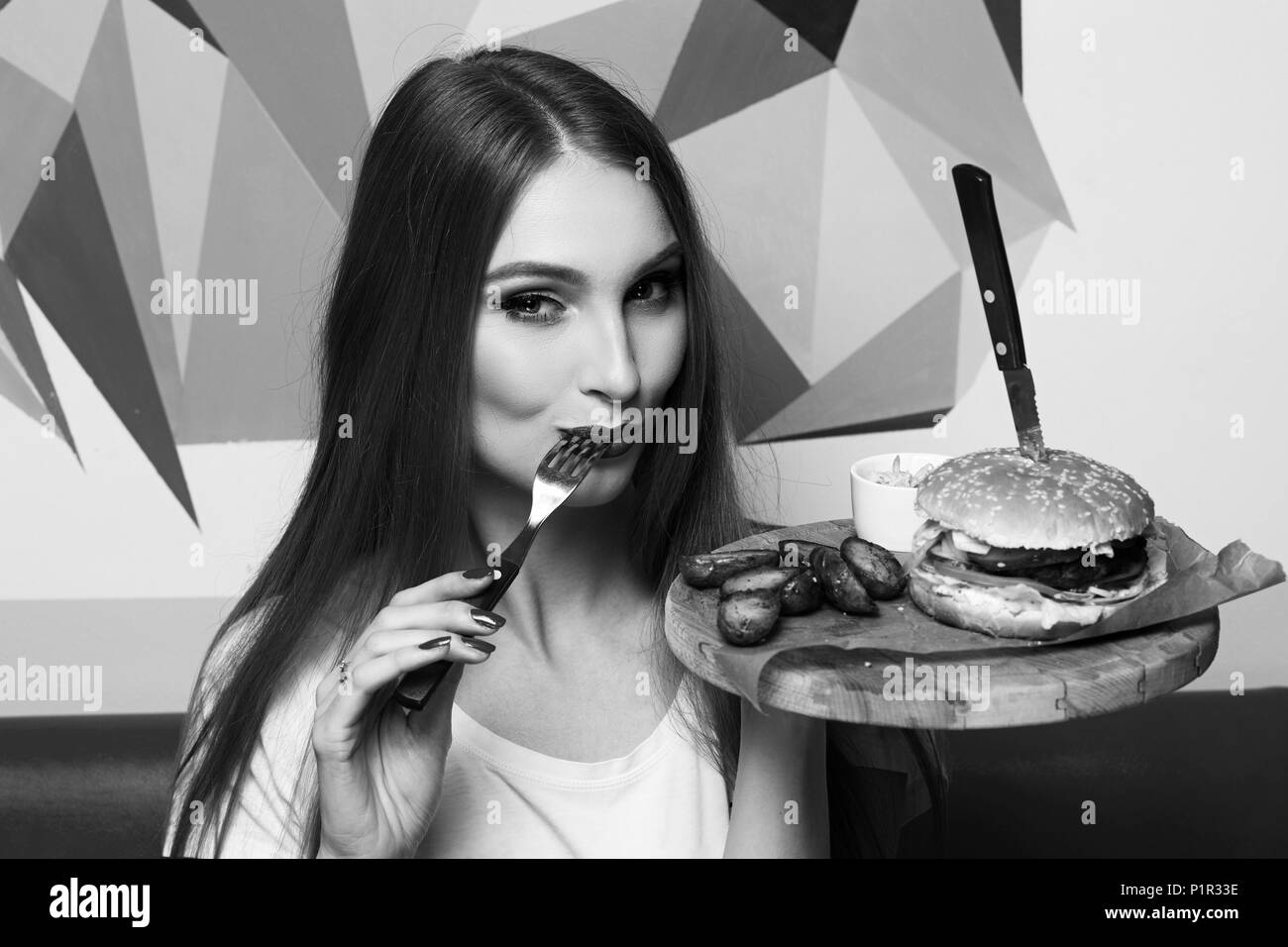 Happy attractive woman holding fourchette et table ronde en bois avec plaque d'appétissants burger et frites. Belle femme joyeuse model eating cheeseburger. Banque D'Images