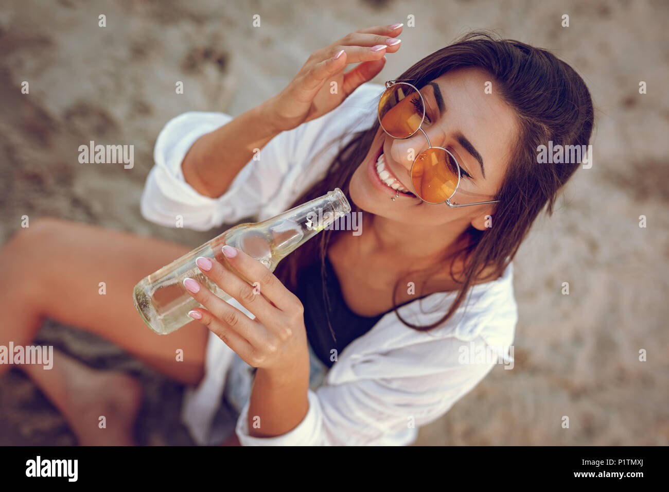 Jeune femme avec des lunettes de détente à l'heure du coucher du soleil sur la plage. Elle est assise au bord de la rivière et de boire de la bière. Vue d'en haut. Banque D'Images