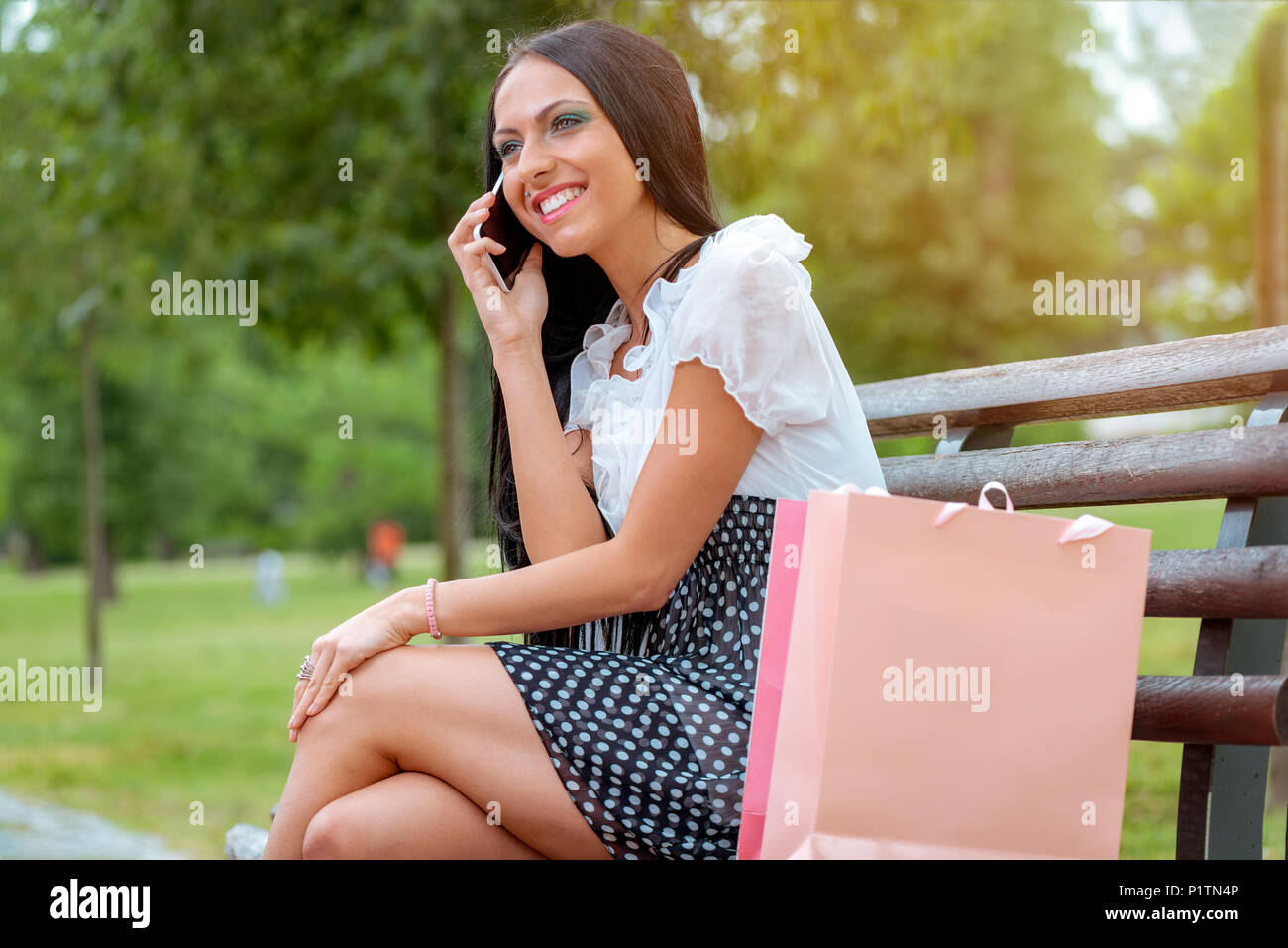 Cheerful girl après shopping avec de nombreux sacs shopping assis sur un banc dans le parc et de parler au téléphone intelligent. Banque D'Images