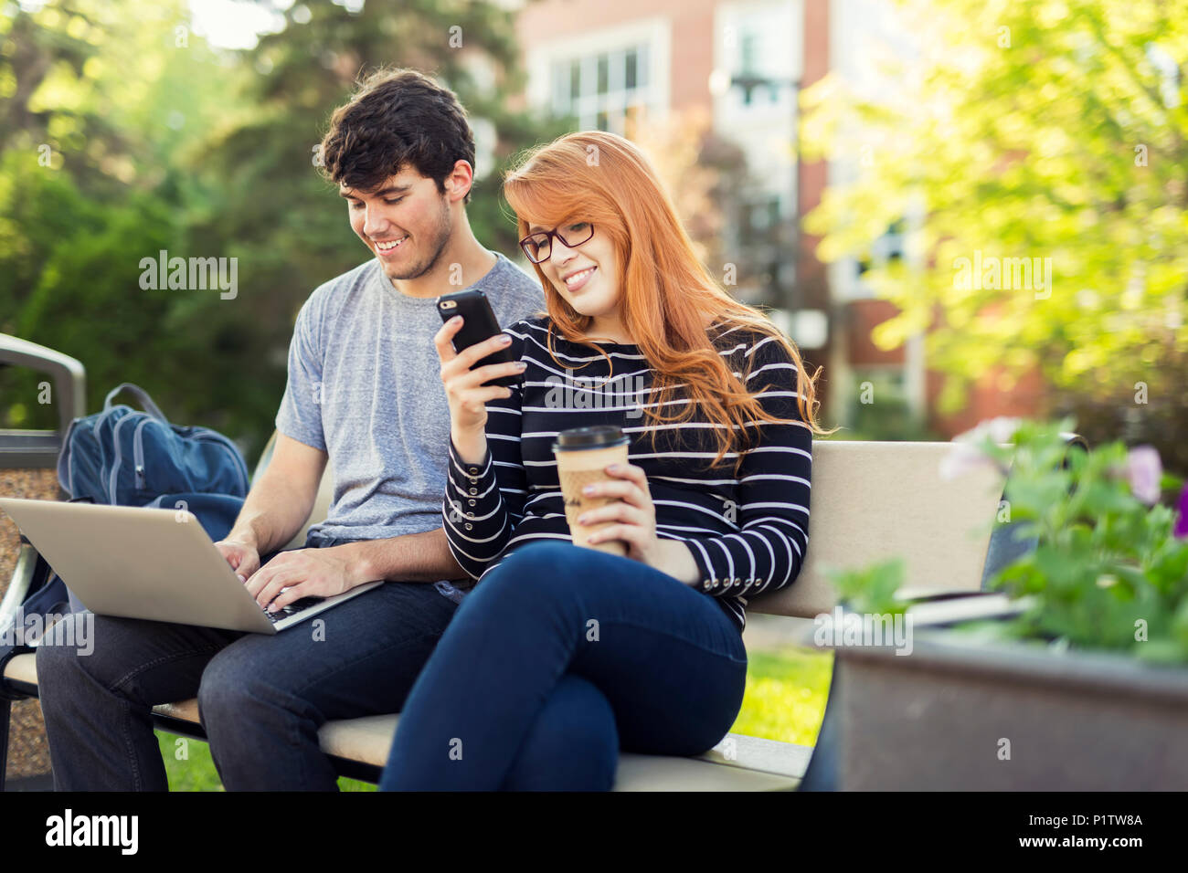 Un jeune homme et une jeune femme s'assoient sur un banc sur le campus universitaire à l'aide d'un ordinateur portable et vérifient les médias sociaux sur un smartphone tout en... Banque D'Images
