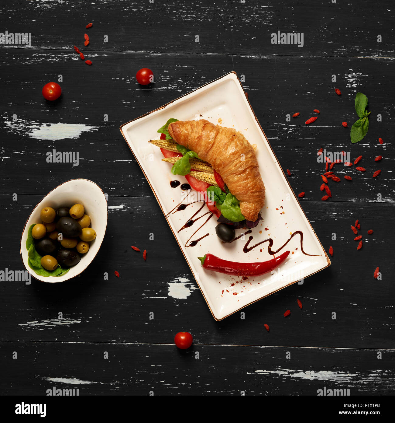 Sandwich appétissant avec des croissants et des légumes sur la plaque rectangulaire plat, bol d'olives et tomates cerises et baies de goji séchées sc Banque D'Images