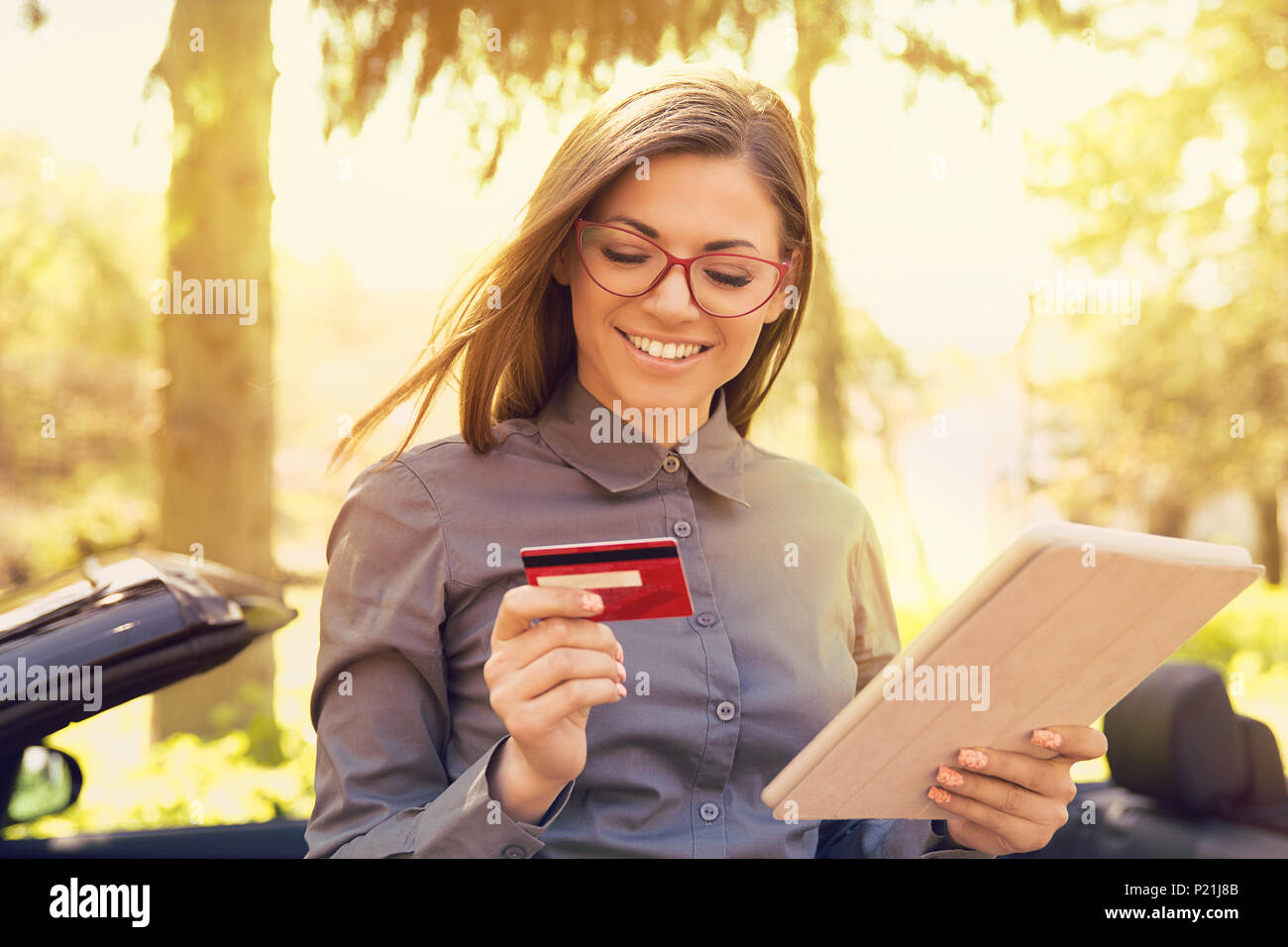 Smiling woman standing par sa nouvelle voiture faire le paiement en ligne sur son ordinateur tablette à l'extérieur sur une journée ensoleillée Banque D'Images