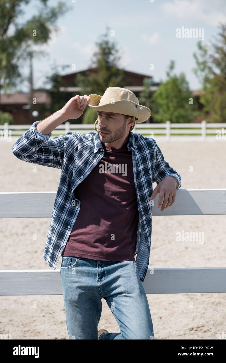 Handsome man wearing cowboy hat et chemise à carreaux leaning on fence au ranch Banque D'Images