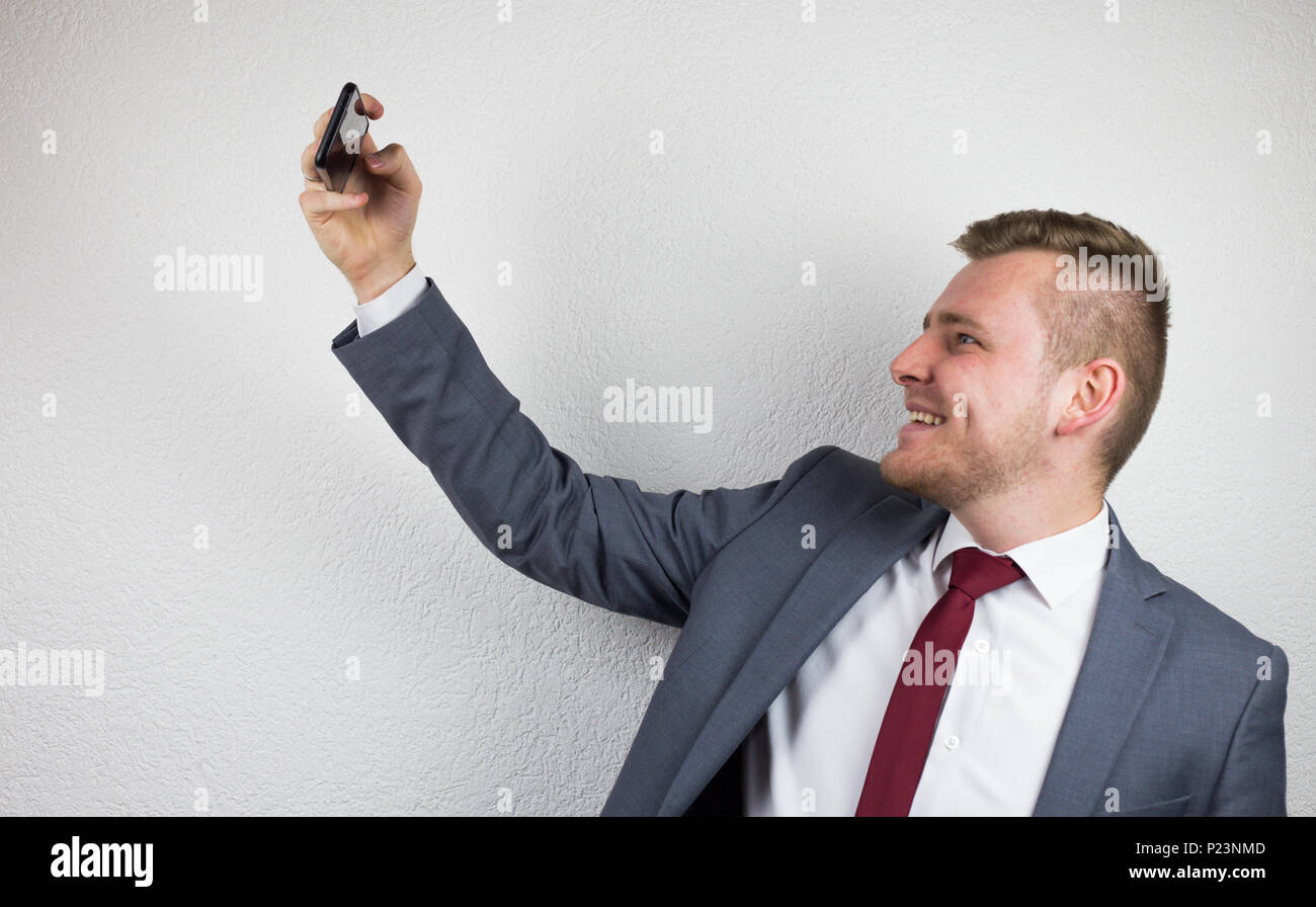 Un jeune homme en costume, à l'aide d'une caméra avant d'un téléphone mobile, un sourire, un fond blanc, un studio Banque D'Images