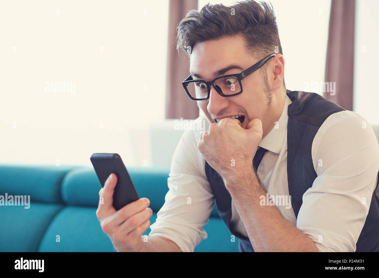 Jeune homme dans l'appartement regardant des smartphone et de mordre dans l'expression de la peur le poing. Banque D'Images