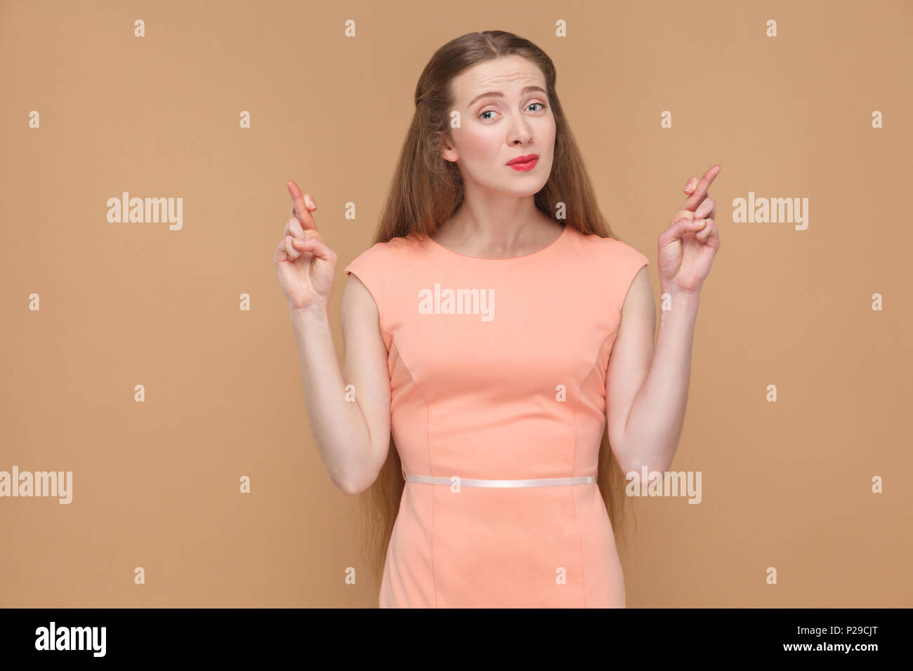 Oh s'il vous plaît. espoir femme avec les doigts croisés looking at camera. mignon, jolie femme avec le maquillage et les cheveux longs en robe rose, studio shot, est Banque D'Images