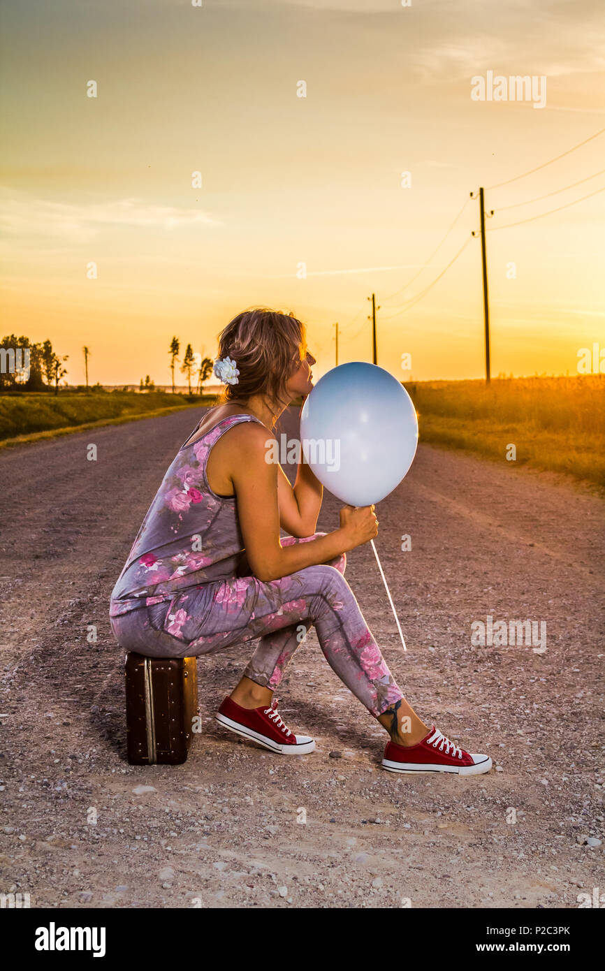 Femme assise sur une vieille valise au milieu d'une route rurale, avec un ballon bleu dans sa main Banque D'Images