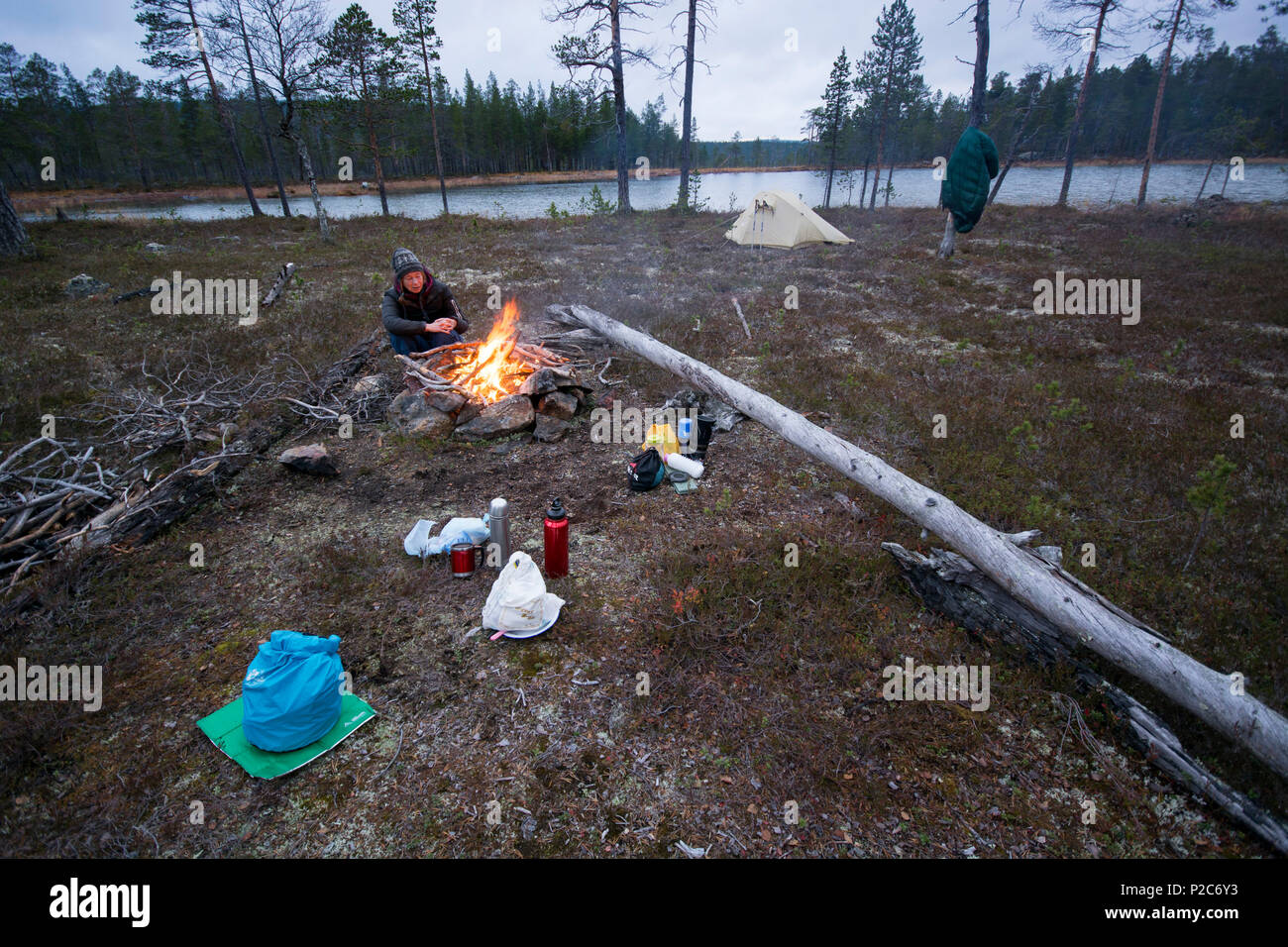 Une jeune femme portant une veste et un chapeau bas assis devant un feu de camp dans une forêt clairsemée, en face de son matériel de bivouac Banque D'Images