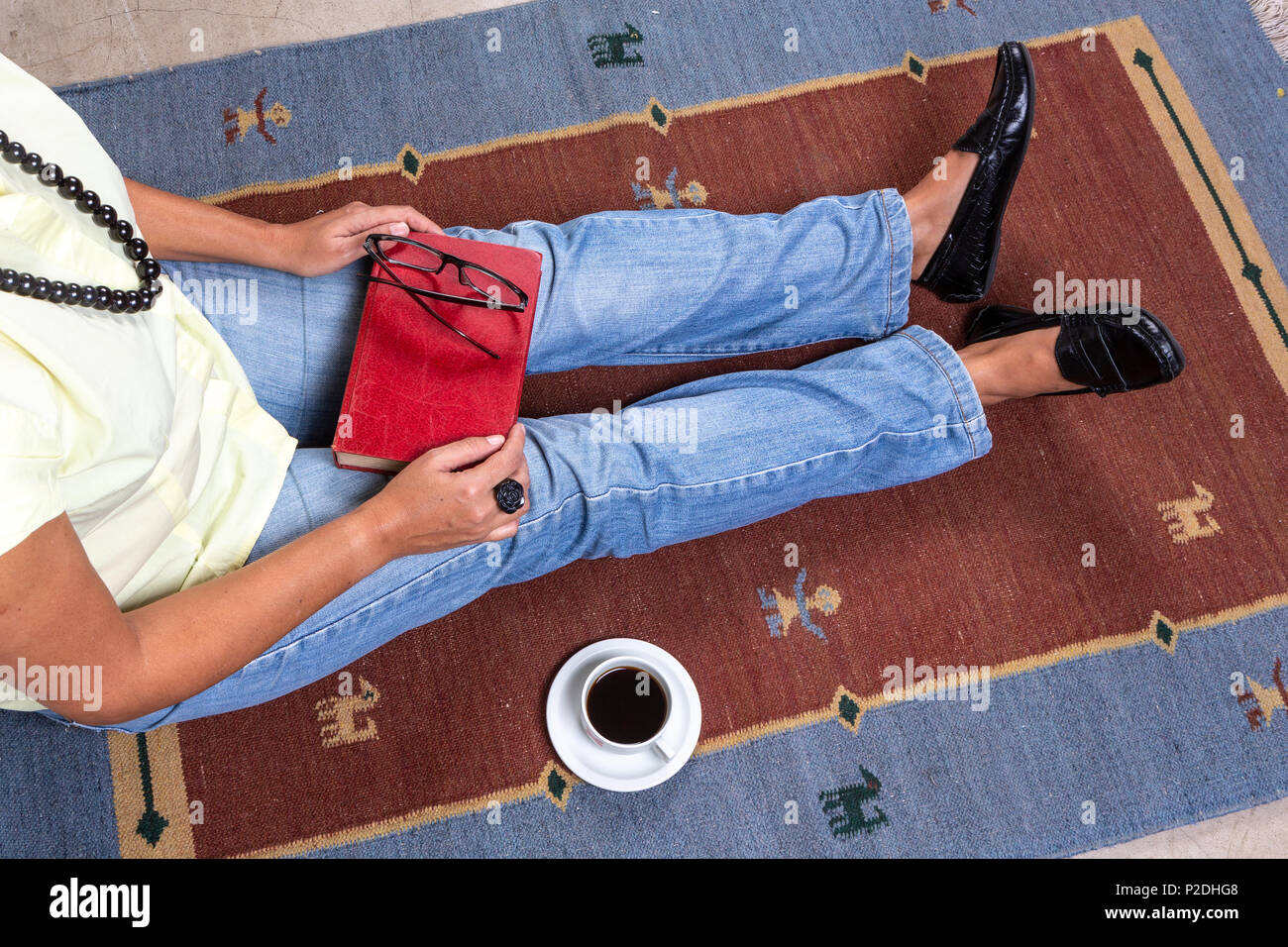 Portrait de jeune femme portant des vêtements décontractés et collier assis sur les tapis, tenant un livre vintage rouge, une tasse de café, et lunettes noires. Banque D'Images
