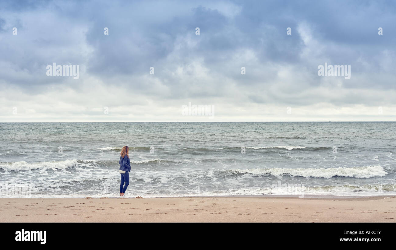 Femme marche le long du bord du surf sur une plage dans un costume de denim bleu à la mer par une froide journée nuageuse with copy space Banque D'Images