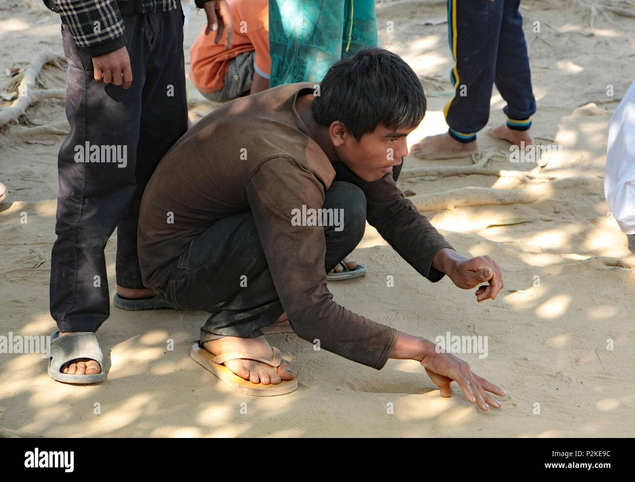 Un garçon cambodgien s'accroupit sur le sol de la lecture d'un jeu de billes se concentrer comme il chiquenaudes la balle en jeu. Dans l'aire de jeux de l'École de rêve. Banque D'Images