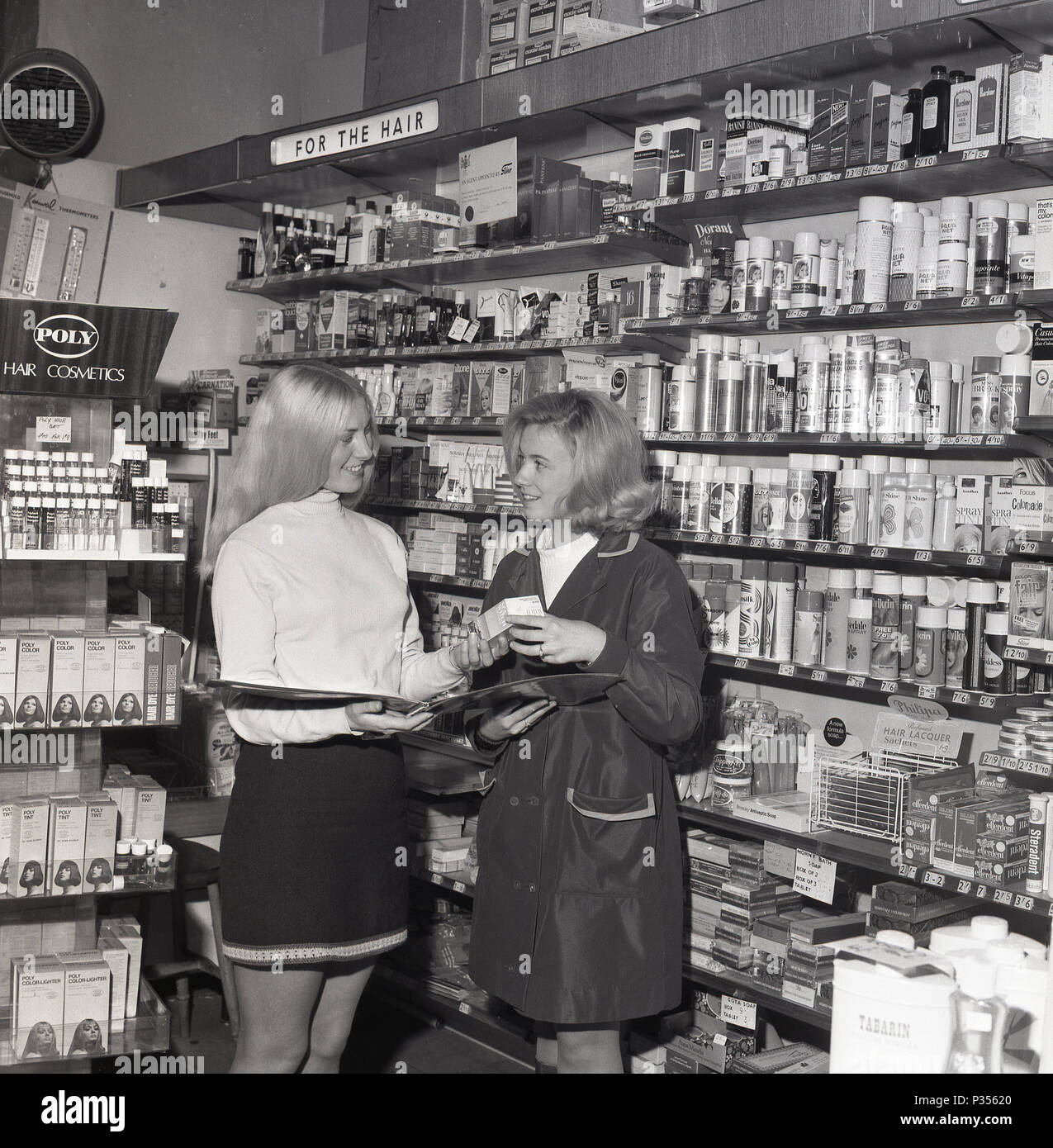 1970, deux jeunes femmes dans une pharmacie, une avec les cheveux longs et portait une jupe courte, montrant l'autre, dans un rainmac, un échantillon de la large gamme de produits cosmétiques et de cheveux disponibles, England, UK. Banque D'Images
