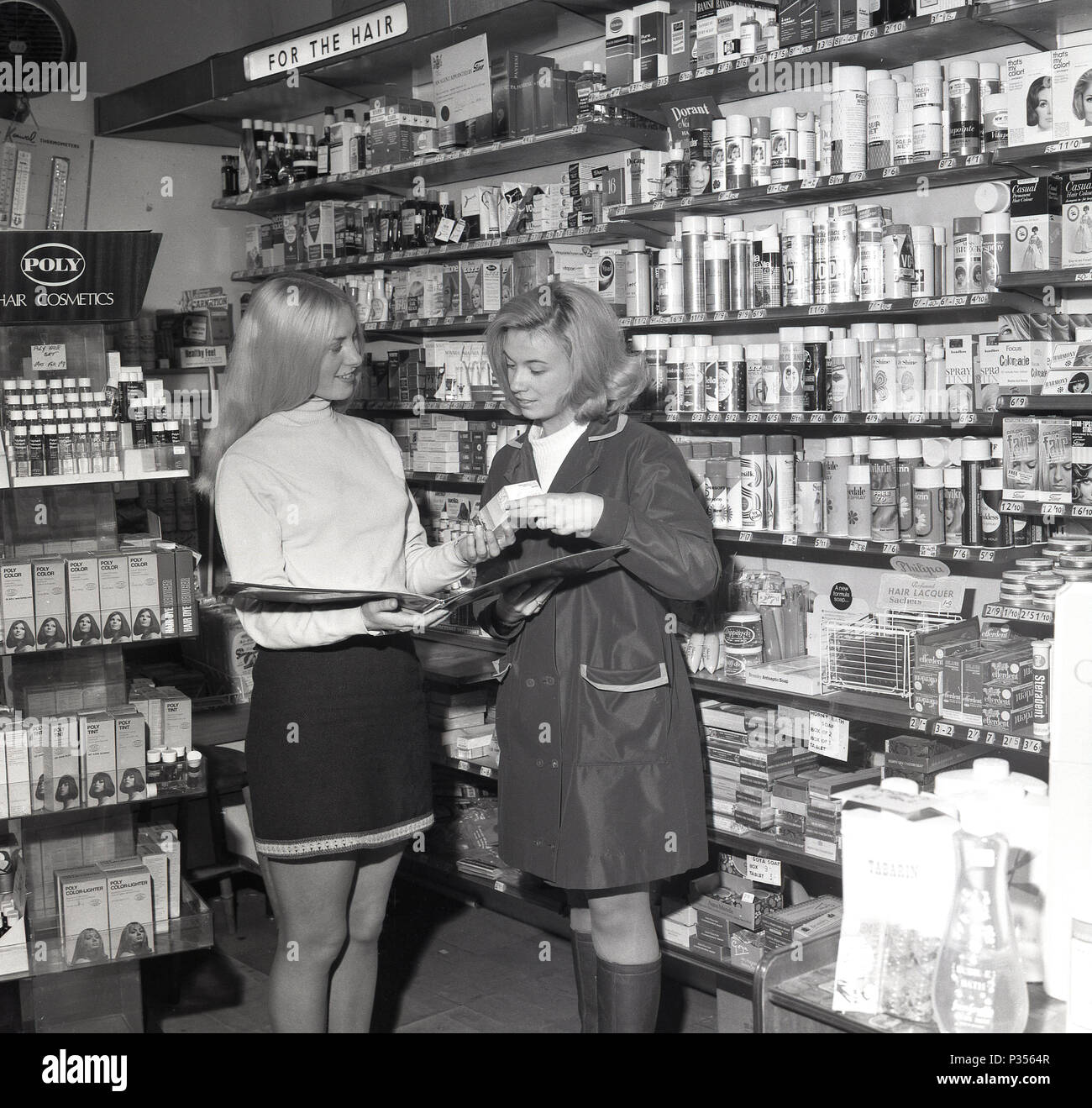 1970, deux jeunes femmes dans une pharmacie, une avec les cheveux longs et portait une jupe courte, montrant l'autre, dans un rainmac, la gamme de cosmétiques et de produits de cheveux disponibles, England, UK. Banque D'Images