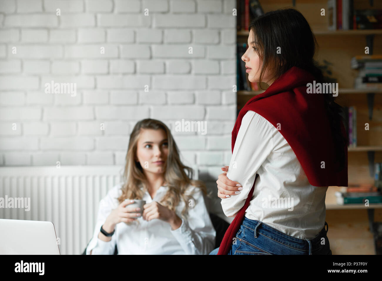 Deux jeunes amis féminins portant des chemises blanches classique décontracté communiquer,travailler avec ordinateur portable, assis dans cette chambre spacieuse avec des murs blancs. Boire du café, de l'apprentissage. D'affaires, des pigistes. Banque D'Images