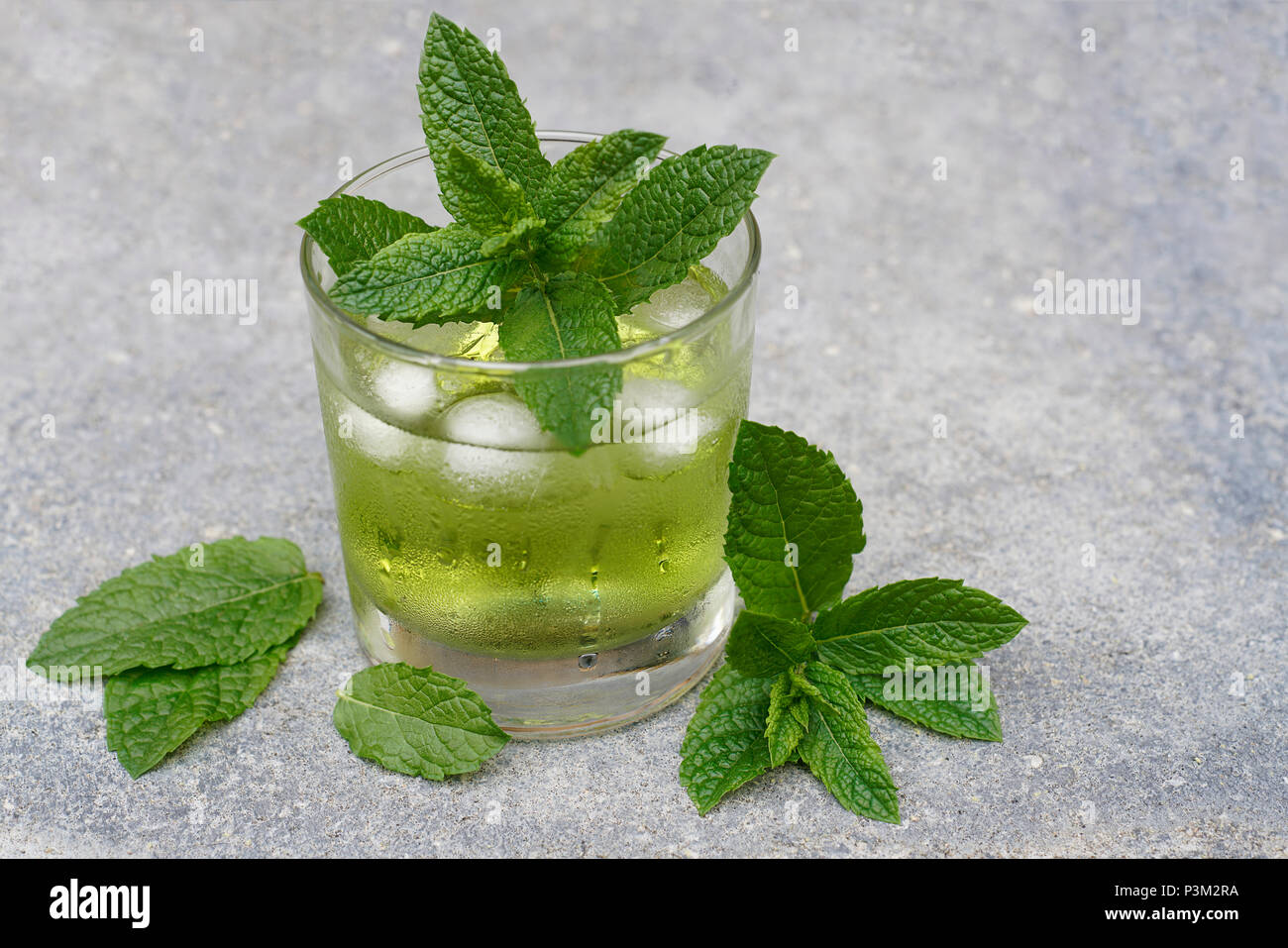 Un verre de liquide vert clair est refroidi avec des glaçons et de la buée. Il se dresse sur un sol en béton. Les feuilles de menthe de l'herbe "menthe maroc' decorat Banque D'Images