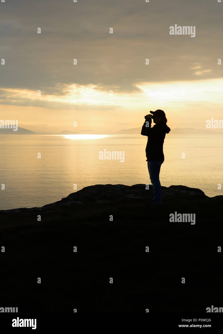La silhouette d'une femme à l'aide de jumelles pour observer la faune au coucher du soleil le long de la côte à Neist Point sur l'île de Skye, Écosse, Royaume-Uni Banque D'Images